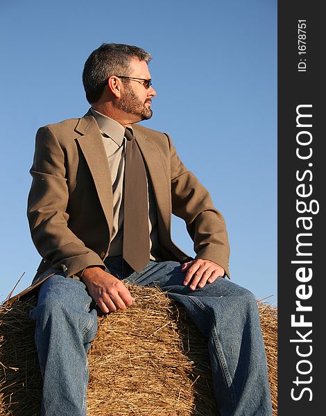 Man in jacket, tie and jeans sitting on a haystack.