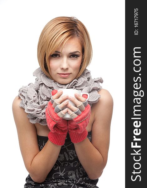 A frontal portrait of a young, blonde woman, holding a mug with both her hands, wearing a muffler and winter gloves, with an expression of being cold. A frontal portrait of a young, blonde woman, holding a mug with both her hands, wearing a muffler and winter gloves, with an expression of being cold.