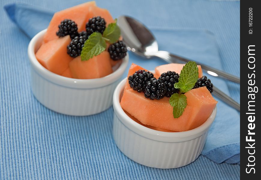 Fresh cantaloupe and blackberries in white ramekins on blue breakfast linen. Selective focus on front bowl. Fresh cantaloupe and blackberries in white ramekins on blue breakfast linen. Selective focus on front bowl.