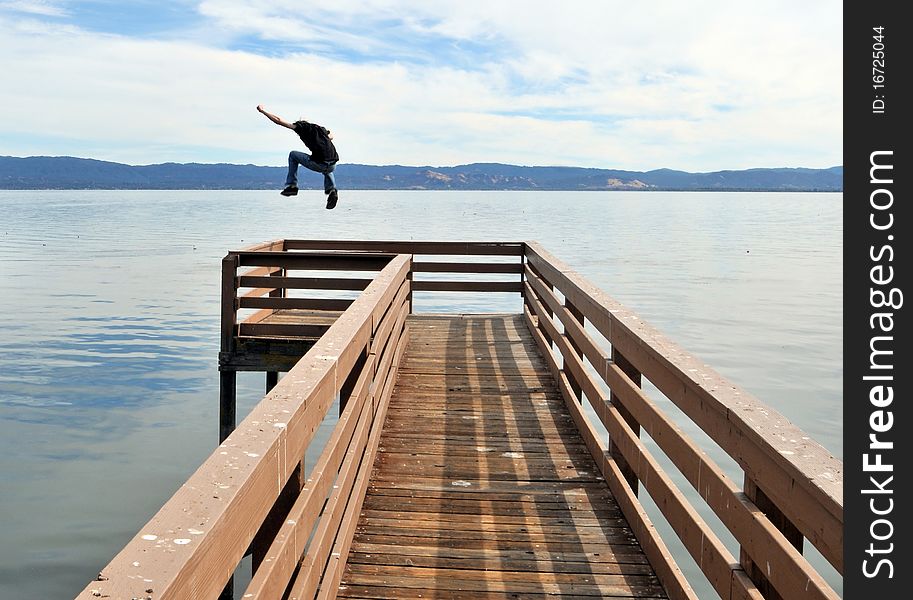 A man jumping a long narrow wooden pier into the calm blue ocean water with outstretched victorious arms. A man jumping a long narrow wooden pier into the calm blue ocean water with outstretched victorious arms.