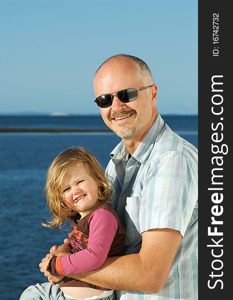 Portrait of mature father holding his toddler girl in front of the blue ocean on a sunny day. Portrait of mature father holding his toddler girl in front of the blue ocean on a sunny day
