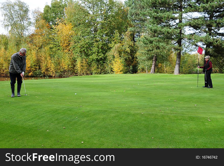 Male golfer preparing to put on the green while a female golfer is holding the putting flag. Male golfer preparing to put on the green while a female golfer is holding the putting flag