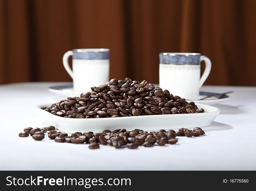 Plate full of roasted arabica coffee beans with two cups of coffee in the background. Plate full of roasted arabica coffee beans with two cups of coffee in the background