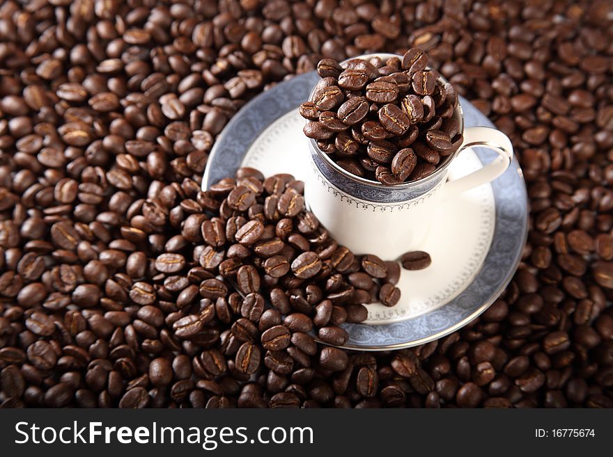 Cup and saucer filled to the top with roasted coffee beans placed on a background of coffee beans. Cup and saucer filled to the top with roasted coffee beans placed on a background of coffee beans