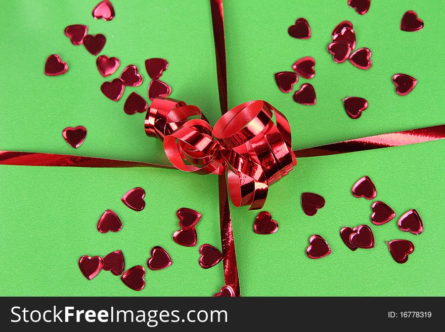 Gift in green wrapping with a red bow decorated with heart shaped confetti in close up. Gift in green wrapping with a red bow decorated with heart shaped confetti in close up