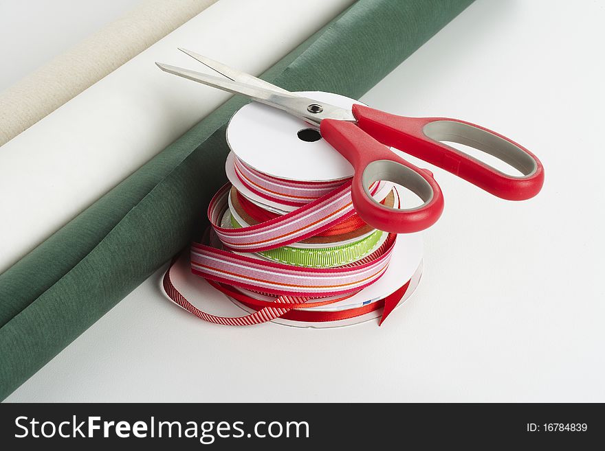 Red handled scissors on top of a stack of red and green ribbon with three rolls of gift wrapping paper. Red handled scissors on top of a stack of red and green ribbon with three rolls of gift wrapping paper