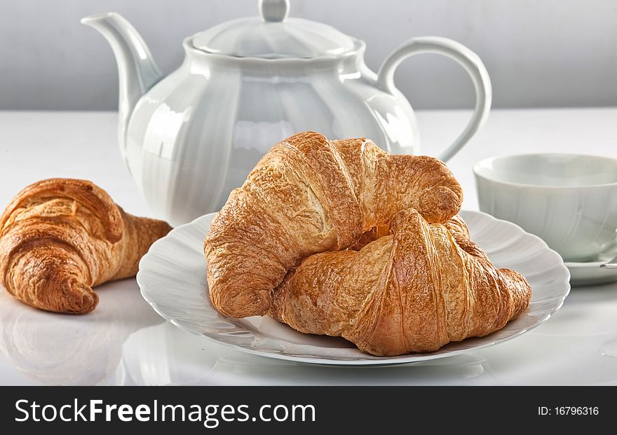 It's time for breakfast! Some fragrant croissant on a white dish. A tea pot is on the background. It's time for breakfast! Some fragrant croissant on a white dish. A tea pot is on the background