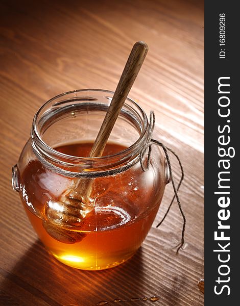 Pot of honey and wooden stick in it. Objects are on wooden table.