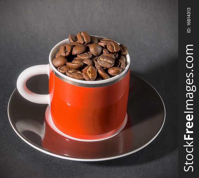 Small Red Cup with Black Saucer filled with Brown Coffee Beans on Grey Background. Small Red Cup with Black Saucer filled with Brown Coffee Beans on Grey Background