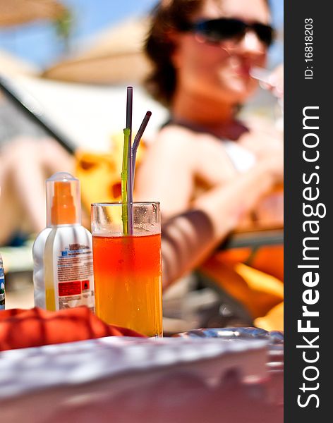 Cocktail glass with two straws, a young women relaxing in the brackground and a sunscreen