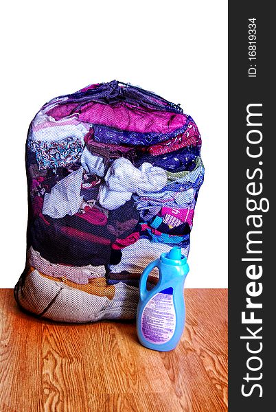 Mesh laundry bag filled with folded clean clothes along with a container of detergent isolated on a white background and wood flooring