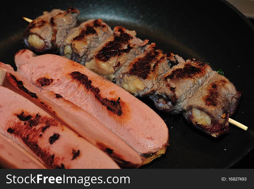 The image depicts a plate of mixed meat, kebabs and wustel during cooking, in a frying pan.