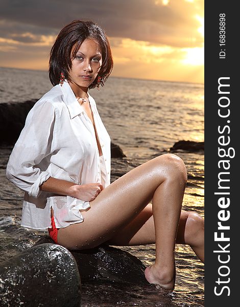 Stunning young girl wearing orange bikini bottoms and white shirt, sitting on the rocks with feet in sea. Late afternoon sunset sky in background. Stunning young girl wearing orange bikini bottoms and white shirt, sitting on the rocks with feet in sea. Late afternoon sunset sky in background.