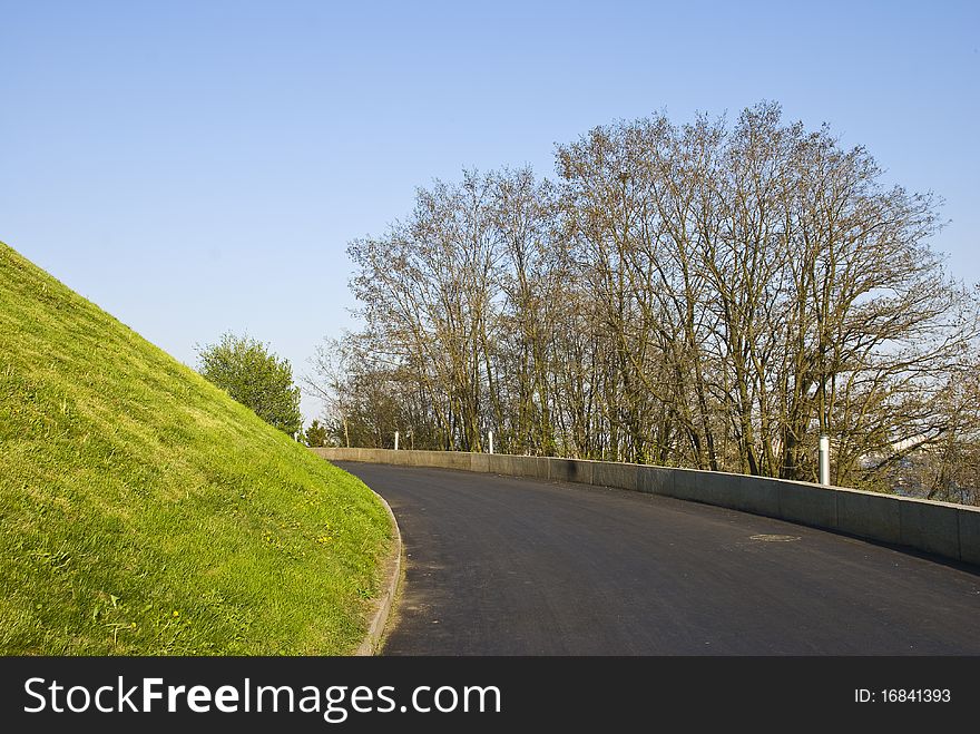 Road with a black coating in the park, the envelope of a hill with a growing spring grass on it. on the other side of the road there are trees and shrubs. Road with a black coating in the park, the envelope of a hill with a growing spring grass on it. on the other side of the road there are trees and shrubs