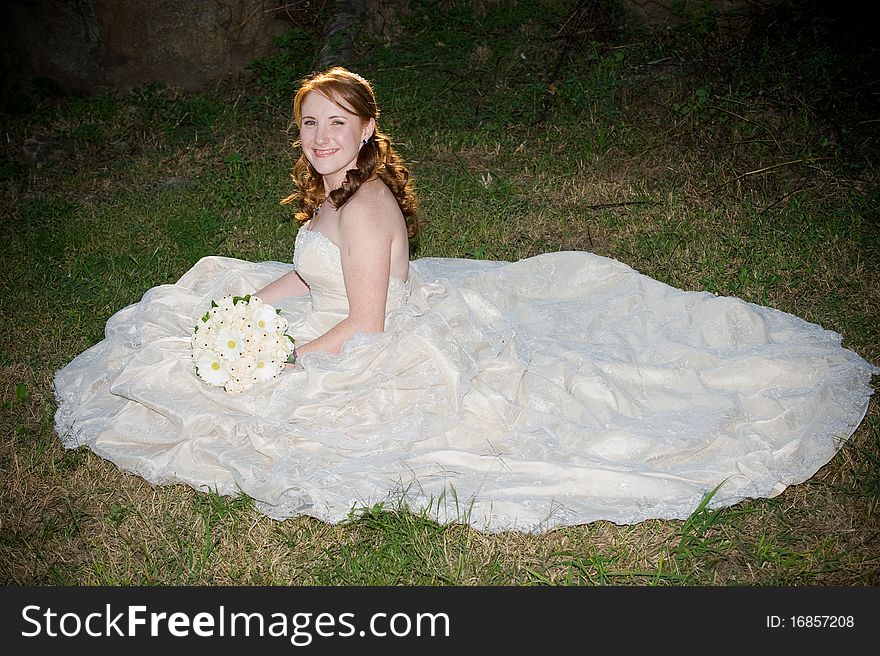 Red head beautiful bride in white dress and bouquet sitting and smiling on green grass. Red head beautiful bride in white dress and bouquet sitting and smiling on green grass