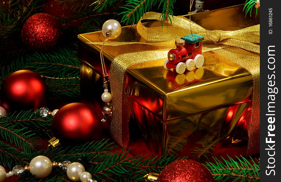 A gold box tied with gold ribbon surrounded by red christmas toys and christmas tree. A gold box tied with gold ribbon surrounded by red christmas toys and christmas tree