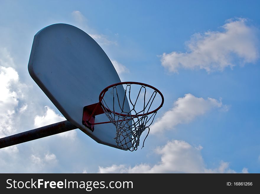 An empty basketball hoop, net swaying in the breeze, against a bright blue sky, with large clouds drifting from behind the backboard. An empty basketball hoop, net swaying in the breeze, against a bright blue sky, with large clouds drifting from behind the backboard.