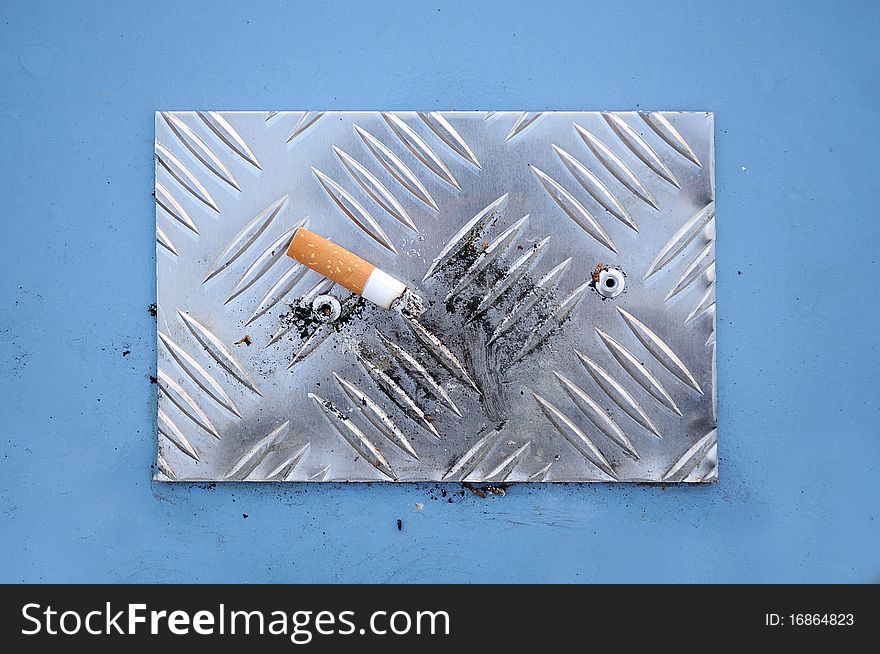 A cigarette end with ash on a metal cigarette stubbing plate on top of a litter bin. A cigarette end with ash on a metal cigarette stubbing plate on top of a litter bin