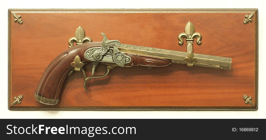 Photo of a Decorative Flintlock Pistol, attached on a wooden, rectangle shaped pedestal. There are four fleur de lis emblems in the corners.