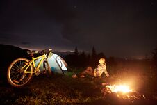 Woman Resting At Night Camping Near Campfire, Tourist Tent, Bicycle Under Evening Sky Full Of Stars Royalty Free Stock Photography