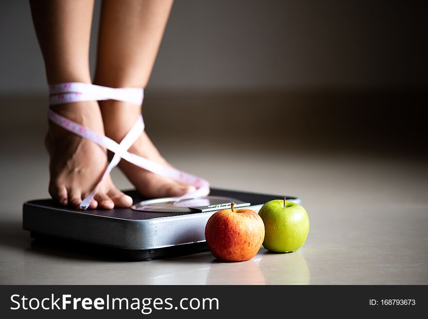 Female leg stepping on weigh scales with measuring tape and green apple. Healthy lifestyle, food and sport concept
