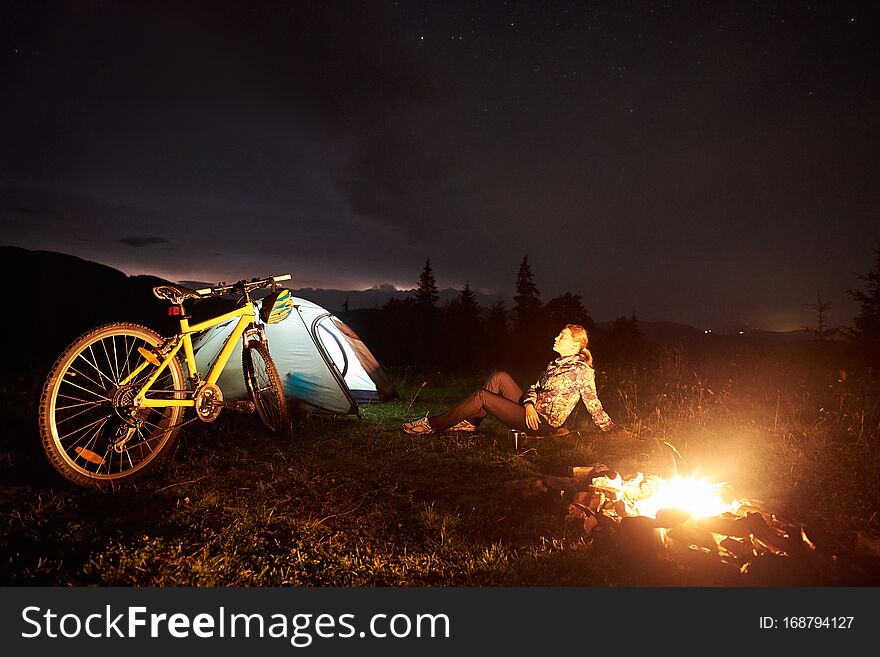 Young woman cyclist having a rest at night camping near burning campfire, illuminated tourist tent, mountain bike under beautiful evening sky full of stars. Outdoor activity and tourism concept. Young woman cyclist having a rest at night camping near burning campfire, illuminated tourist tent, mountain bike under beautiful evening sky full of stars. Outdoor activity and tourism concept