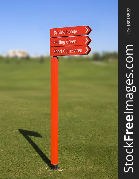 A red pole with sign arrows, pointing directions in a golf course: Driving Range, Putting Green, Short Game Area, over a clear blue sky with a blurred fairway on background. A red pole with sign arrows, pointing directions in a golf course: Driving Range, Putting Green, Short Game Area, over a clear blue sky with a blurred fairway on background