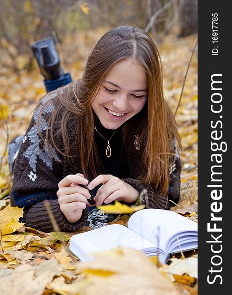 Portrait of smiling brown-haired girl in autumn park laying on leaves, reading a book. Portrait of smiling brown-haired girl in autumn park laying on leaves, reading a book