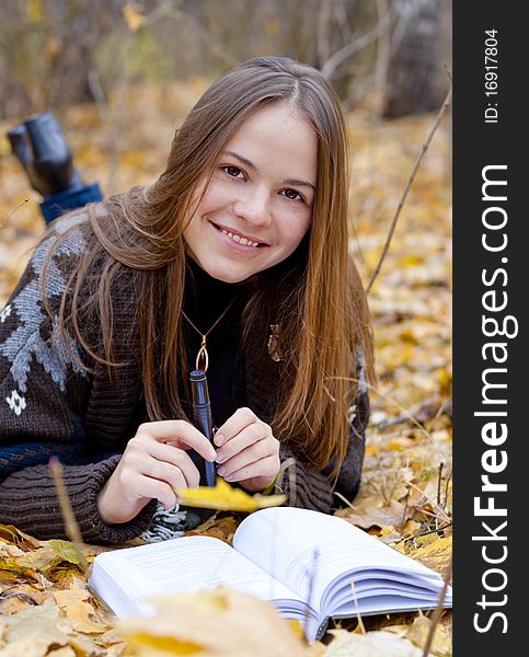 Portrait of smiling brown-haired girl in autumn park laying on leaves, reading a book. Portrait of smiling brown-haired girl in autumn park laying on leaves, reading a book