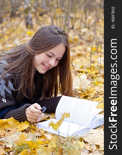 Portrait of smiling brown-haired girl in autumn park laying on leaves, reading a book. Portrait of smiling brown-haired girl in autumn park laying on leaves, reading a book