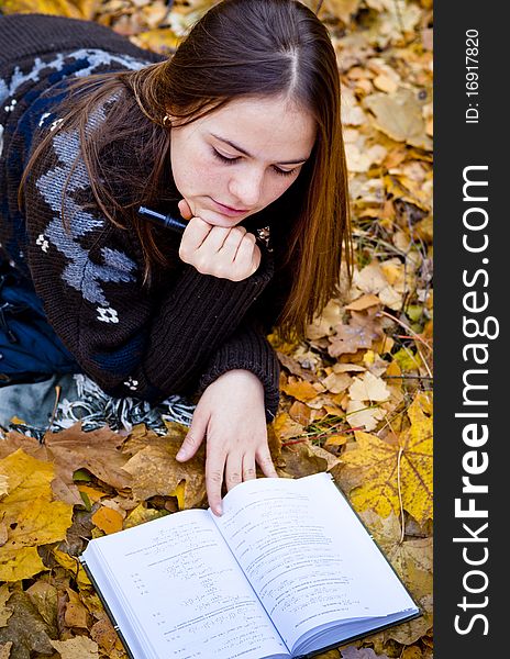 Portrait of thoughtful brown-haired girl in autumn park laying on leaves, reading a book. Portrait of thoughtful brown-haired girl in autumn park laying on leaves, reading a book