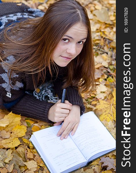 Portrait of brown-haired girl in autumn park laying on leaves, reading a book