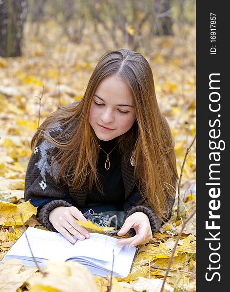 Portrait of brown-haired girl in autumn park laying on leaves, reading a book