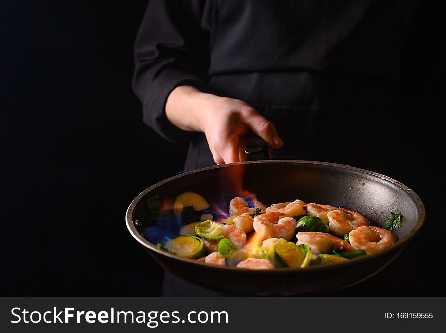 Professional Chef Cook On A Dark Background Prepares Seafood, Shrimp With Fire And Vegetables. Roasting And Cooking Veggie Food.