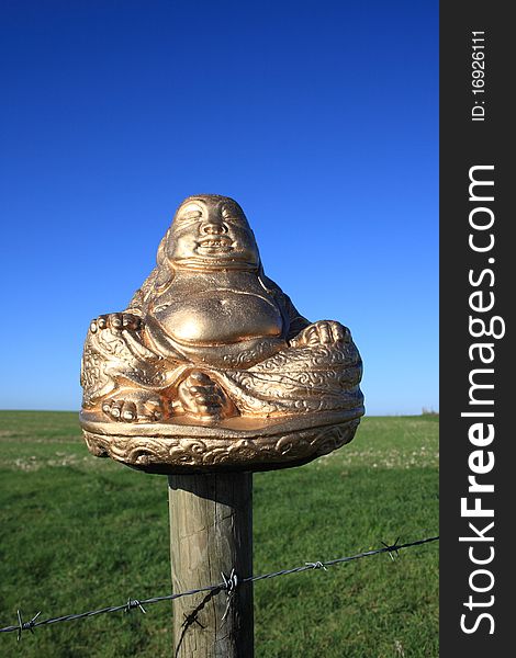 A beautiful golden Buddha sitting on a barbed wire fence post with a green field and lovely blue sky behind him. A beautiful golden Buddha sitting on a barbed wire fence post with a green field and lovely blue sky behind him.