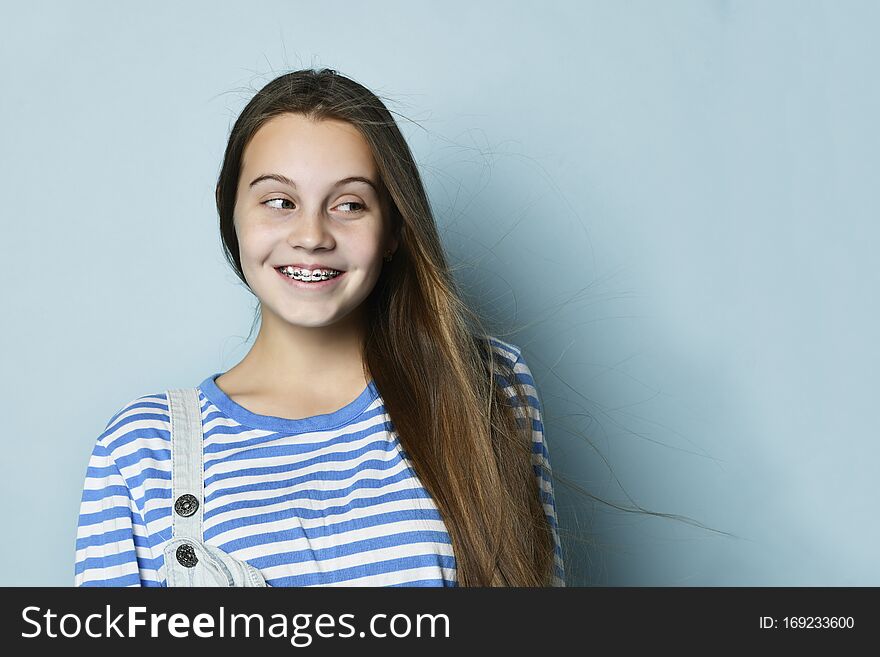 Teen lady in jeans overall and striped sweatshirt. She smiling, looking aside, posing against blue background. Close up