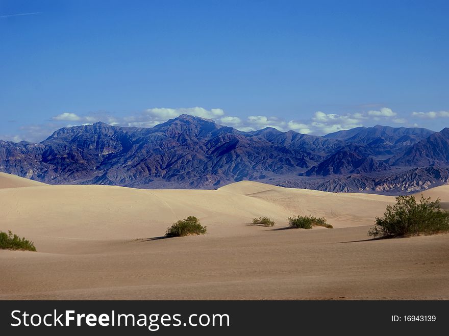 Beautiful, clear photo of Death Valley, California. Sand dunes brush up agains purple mountains with puffy clouds peeking over them. Beautiful, clear photo of Death Valley, California. Sand dunes brush up agains purple mountains with puffy clouds peeking over them.