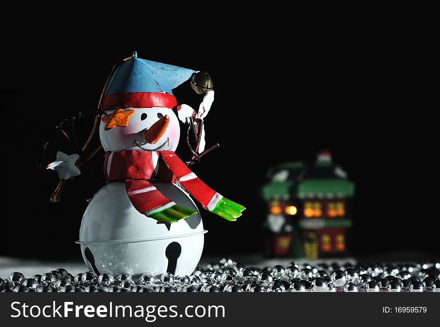 Christmas tree ornament snowman standing among silver balls with black background and lit house. Christmas tree ornament snowman standing among silver balls with black background and lit house