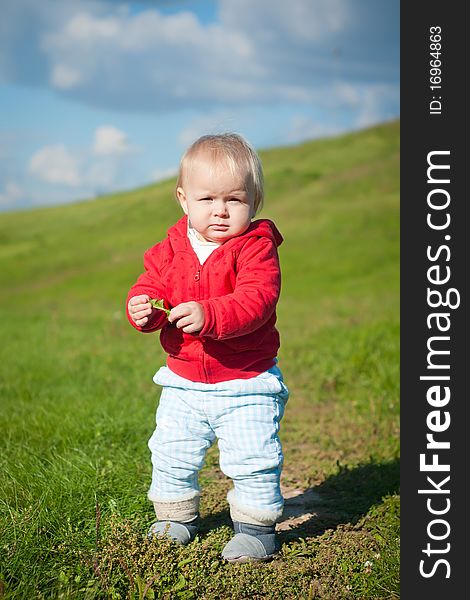 Adorable baby walking through green road within red jacket with hearts on mountain side. holding grass and looking forward. Adorable baby walking through green road within red jacket with hearts on mountain side. holding grass and looking forward