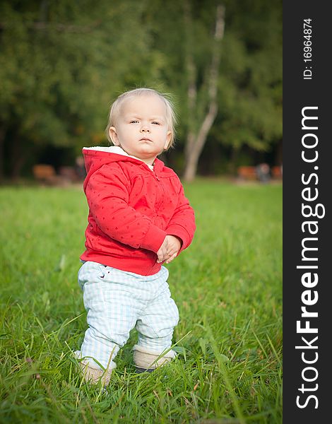 Adorable baby stay on green grass near forest within red jacket with hearts. looking forward