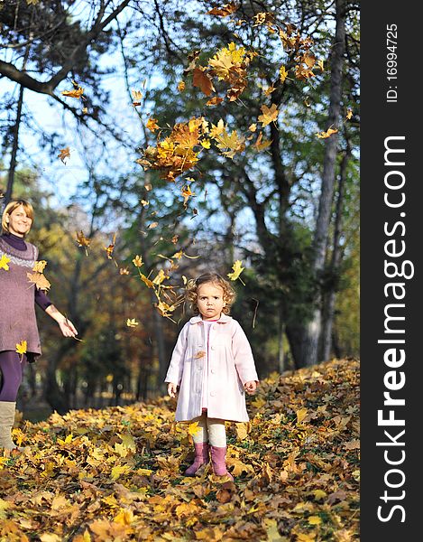 Portrait with little girl in autumn in the park with leaves fallen down on her and mom in the background