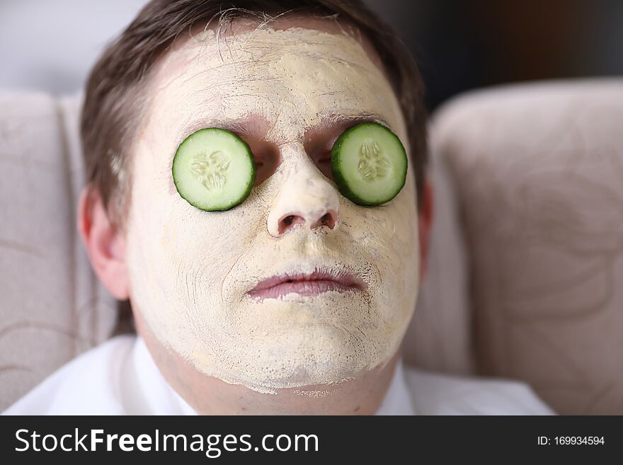 Close-up of young man resting with yellow facial mask. Male person with slices of fresh cucumber on eyes. Guy relaxing during cosmetic procedure. Wellness, skincare and healthcare concept. Close-up of young man resting with yellow facial mask. Male person with slices of fresh cucumber on eyes. Guy relaxing during cosmetic procedure. Wellness, skincare and healthcare concept