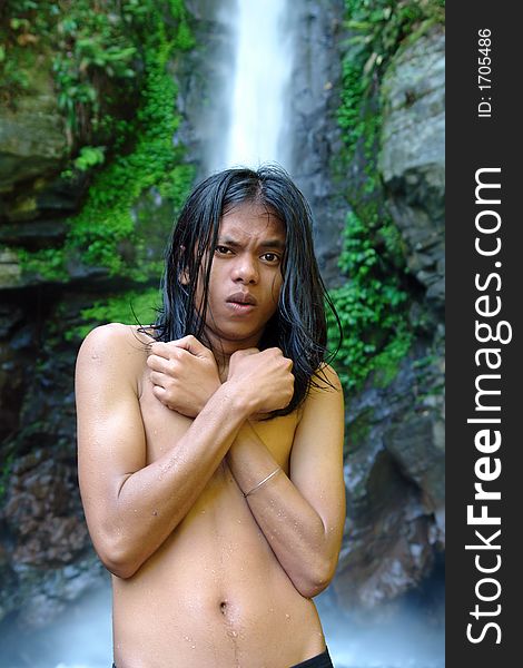 Asian exotic long-haired boy in front of a tropical pristine waterfall, after taking a dip in the whirlpool, shivering and covered with droplets. Asian exotic long-haired boy in front of a tropical pristine waterfall, after taking a dip in the whirlpool, shivering and covered with droplets.