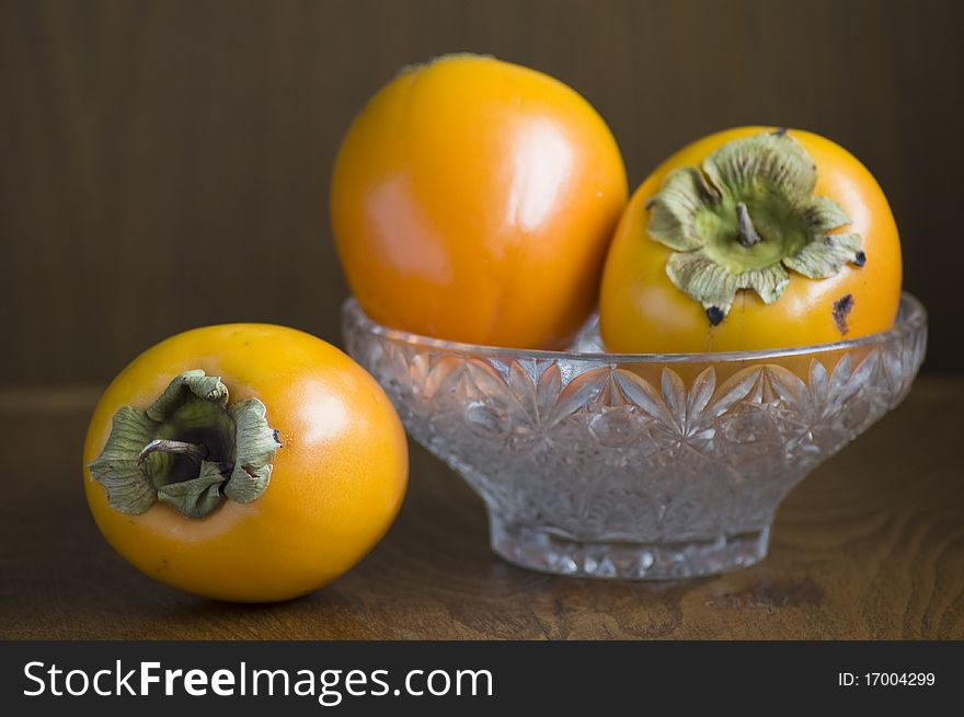 Still life of three Persimmons, two in a glass bowl. Everything decorated in front of dark brown background. Still life of three Persimmons, two in a glass bowl. Everything decorated in front of dark brown background.