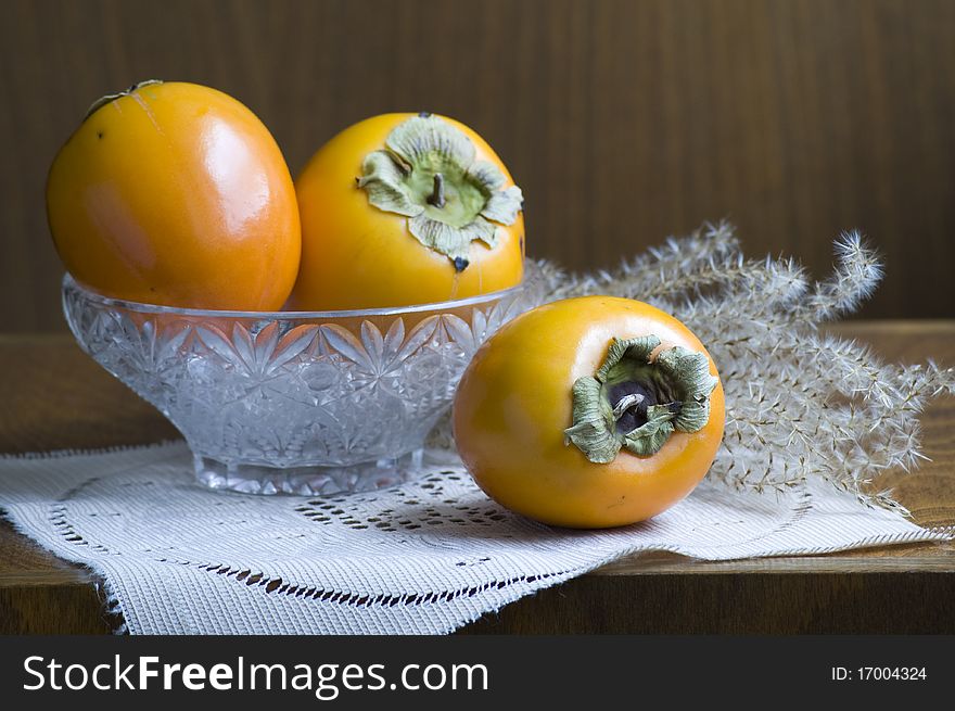 Three persimmons decorated in glass basin in front of dark brown wood background with a autumn grass. Everything is decorated on beige cloth. Three persimmons decorated in glass basin in front of dark brown wood background with a autumn grass. Everything is decorated on beige cloth.