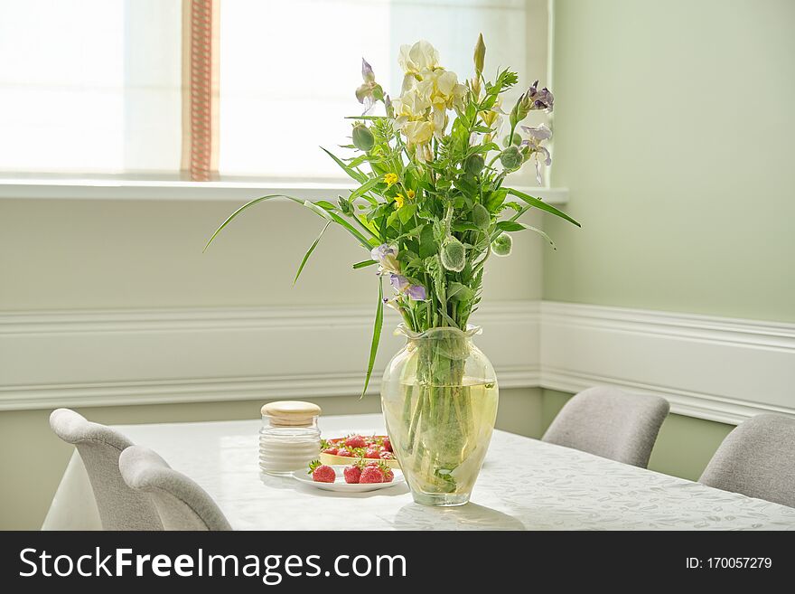 Home dining room interior, table with white tablecloth, spring summer bouquet of flowers in vase, tray with ripe strawberries. Home dining room interior, table with white tablecloth, spring summer bouquet of flowers in vase, tray with ripe strawberries