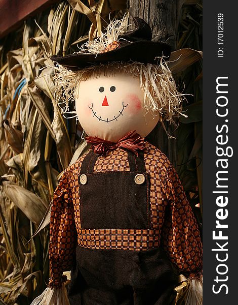 A smiling scarecrow with brown clothes and a floppy hat. There is corn and barnwood in the background. A smiling scarecrow with brown clothes and a floppy hat. There is corn and barnwood in the background