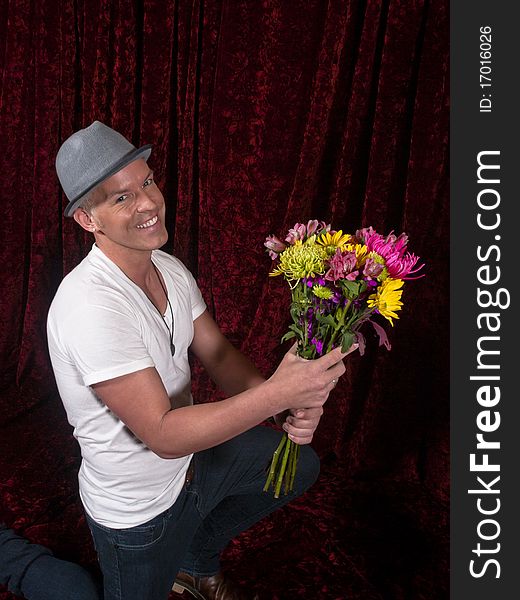 Smiling man wearing hat kneels with bouquet of flowers, photographed against red velvet background. Smiling man wearing hat kneels with bouquet of flowers, photographed against red velvet background