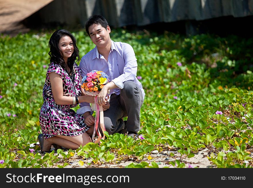 Attractive young couple sitting on beach grass meadow with flower bouquet