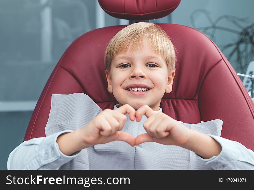 Close Up Portrait Of A Very Happy Boy Sitting In Dentist Chair Showing Heart With His Fingers And Smiling At The Camera, Dental